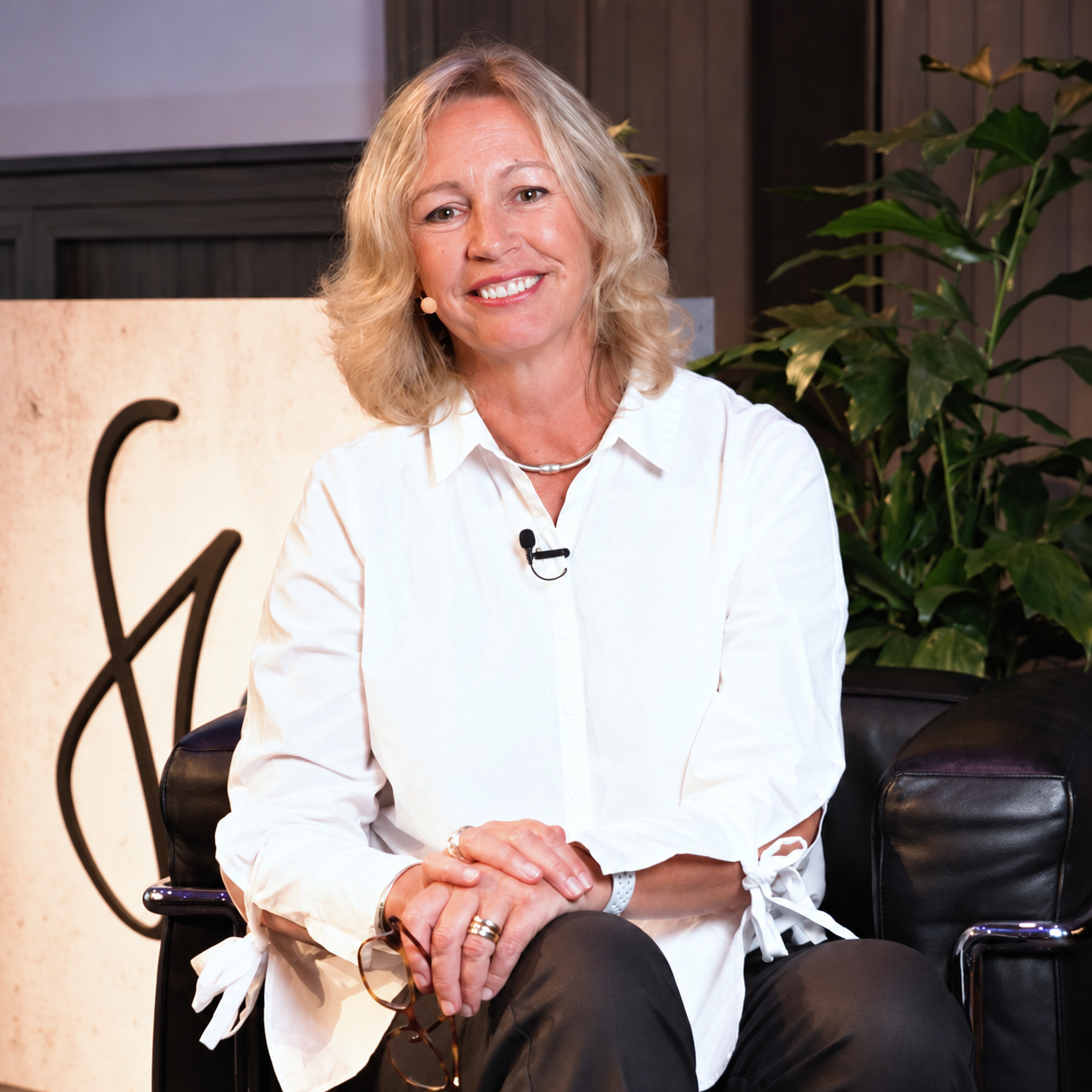 Claudia Brett smiling, seated in a white shirt with her hands resting on her lap.